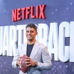 FootballR - NFL - Ein Mann im Anzug hält einen Football in der Hand, während er vor einem Netflix-Schild posiert.