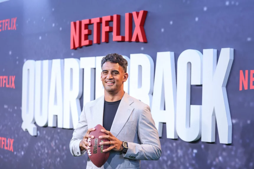 FootballR - NFL - Ein Mann im Anzug hält einen Football in der Hand, während er vor einem Netflix-Schild posiert.