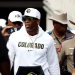 FootballR - NFL - Deion Sanders, bekleidet mit einem weißen Kapuzenpullover, navigiert durch eine Menschenmenge.