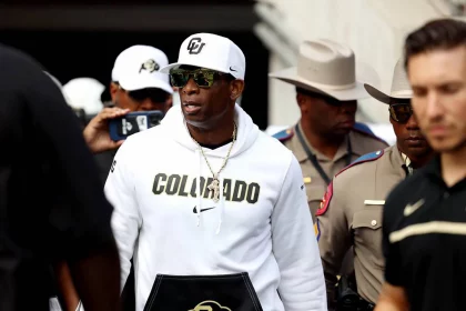 FootballR - NFL - Deion Sanders, bekleidet mit einem weißen Kapuzenpullover, navigiert durch eine Menschenmenge.