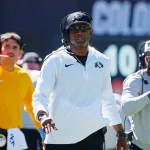 FootballR - Salary Cap - Eine Gruppe Coaches am Spielfeldrand, angeführt von Startrainer Deion Sanders.