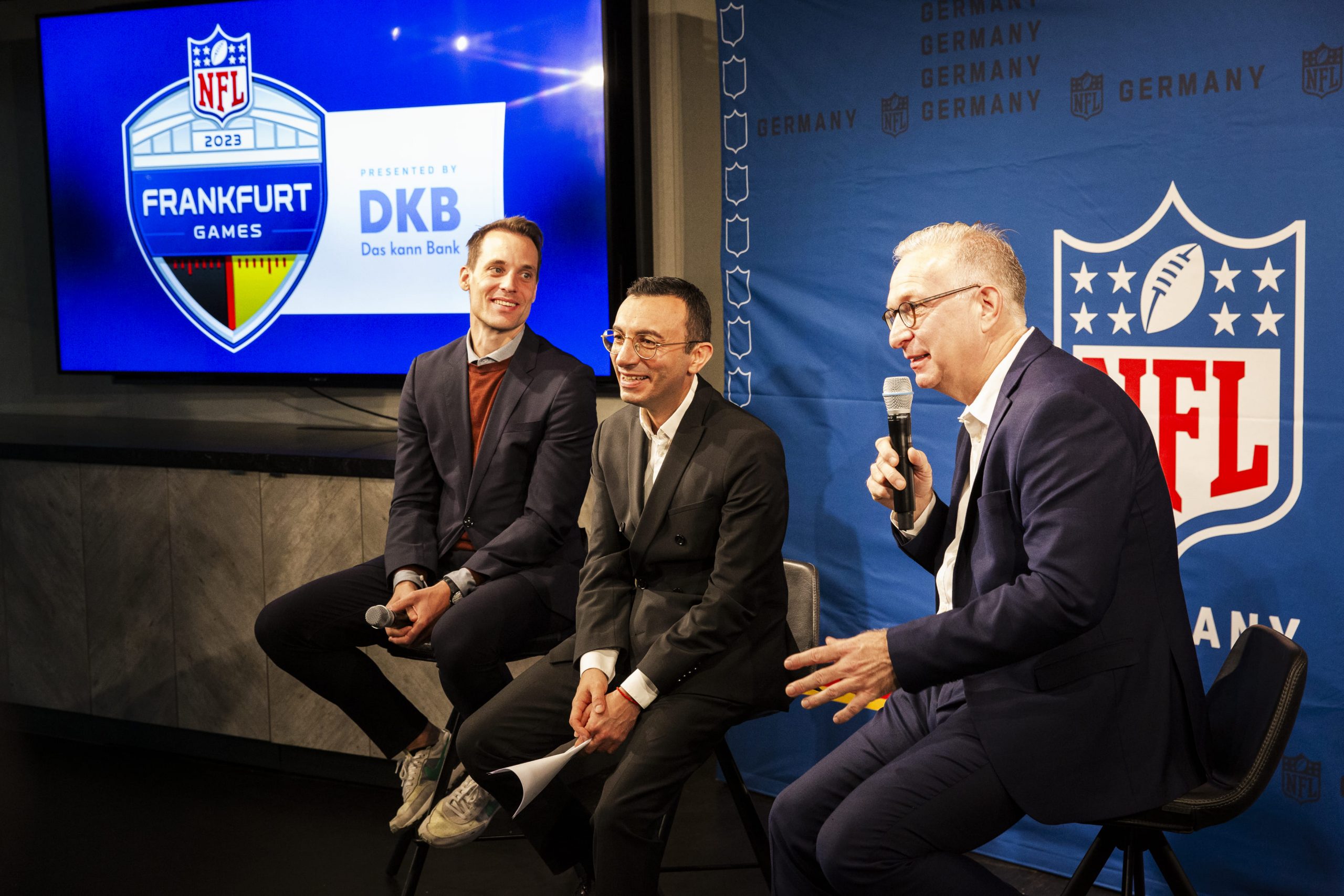 FootballR - NFL - Drei Männer in Anzügen sitzen auf einer Bühne bei einem NFL-Event in Frankfurt, Deutschland.