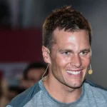 FootballR - NFL - Ein Mann, Tom Brady, lächelt vor einer Menschenmenge bei einer offiziellen Veranstaltung.