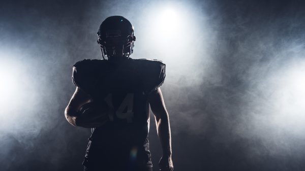 FootballR - NFL - Die Silhouette eines Footballspielers, der einen Ball hält, vor einem dunklen Hintergrund, perfekt, um Ihr DAZN-Abo zu präsentieren.