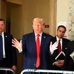 Donald Trump, in marineblauem Anzug und roter Krawatte, gestikuliert mit offenen Händen, während er über die Gesetzesentwürfe ( Traumatische Hirnverletzungen ) von Donald Trump spricht. Ihn flankieren drei ernst wirkende Männer in Anzügen, einer davon mit Aktenordnern. Sie stehen in einem repräsentativen Gebäude mit Marmor- und beigefarbenen Wänden. Ein NYPD-Beamter ist teilweise zu sehen. Diese Beschreibung wurde mit der FootballR KI automatisch generiert.