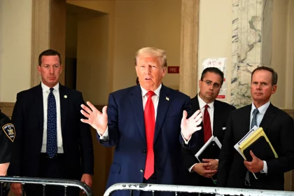 Donald Trump, in marineblauem Anzug und roter Krawatte, gestikuliert mit offenen Händen, während er über die Gesetzesentwürfe ( Traumatische Hirnverletzungen ) von Donald Trump spricht. Ihn flankieren drei ernst wirkende Männer in Anzügen, einer davon mit Aktenordnern. Sie stehen in einem repräsentativen Gebäude mit Marmor- und beigefarbenen Wänden. Ein NYPD-Beamter ist teilweise zu sehen. Diese Beschreibung wurde mit der FootballR KI automatisch generiert.