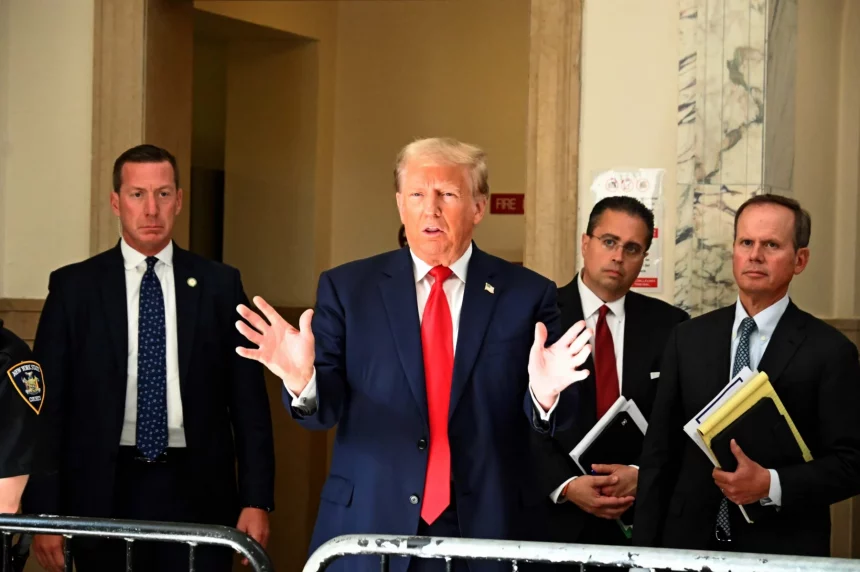 Donald Trump, in marineblauem Anzug und roter Krawatte, gestikuliert mit offenen Händen, während er über die Gesetzesentwürfe ( Traumatische Hirnverletzungen ) von Donald Trump spricht. Ihn flankieren drei ernst wirkende Männer in Anzügen, einer davon mit Aktenordnern. Sie stehen in einem repräsentativen Gebäude mit Marmor- und beigefarbenen Wänden. Ein NYPD-Beamter ist teilweise zu sehen. Diese Beschreibung wurde mit der FootballR KI automatisch generiert.