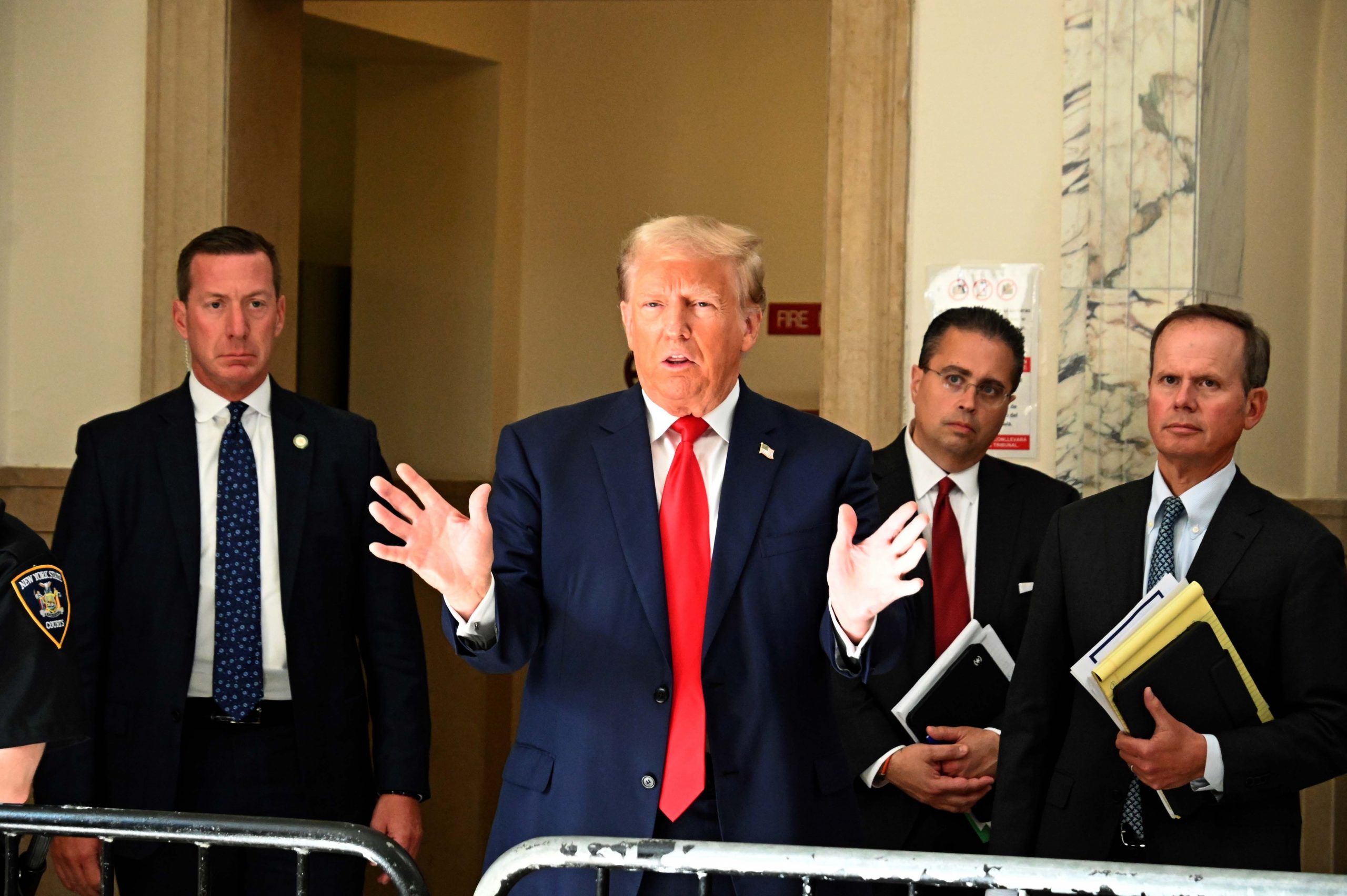 Donald Trump, in marineblauem Anzug und roter Krawatte, gestikuliert mit offenen Händen, während er über die Gesetzesentwürfe ( Traumatische Hirnverletzungen ) von Donald Trump spricht. Ihn flankieren drei ernst wirkende Männer in Anzügen, einer davon mit Aktenordnern. Sie stehen in einem repräsentativen Gebäude mit Marmor- und beigefarbenen Wänden. Ein NYPD-Beamter ist teilweise zu sehen. Diese Beschreibung wurde mit der FootballR KI automatisch generiert.