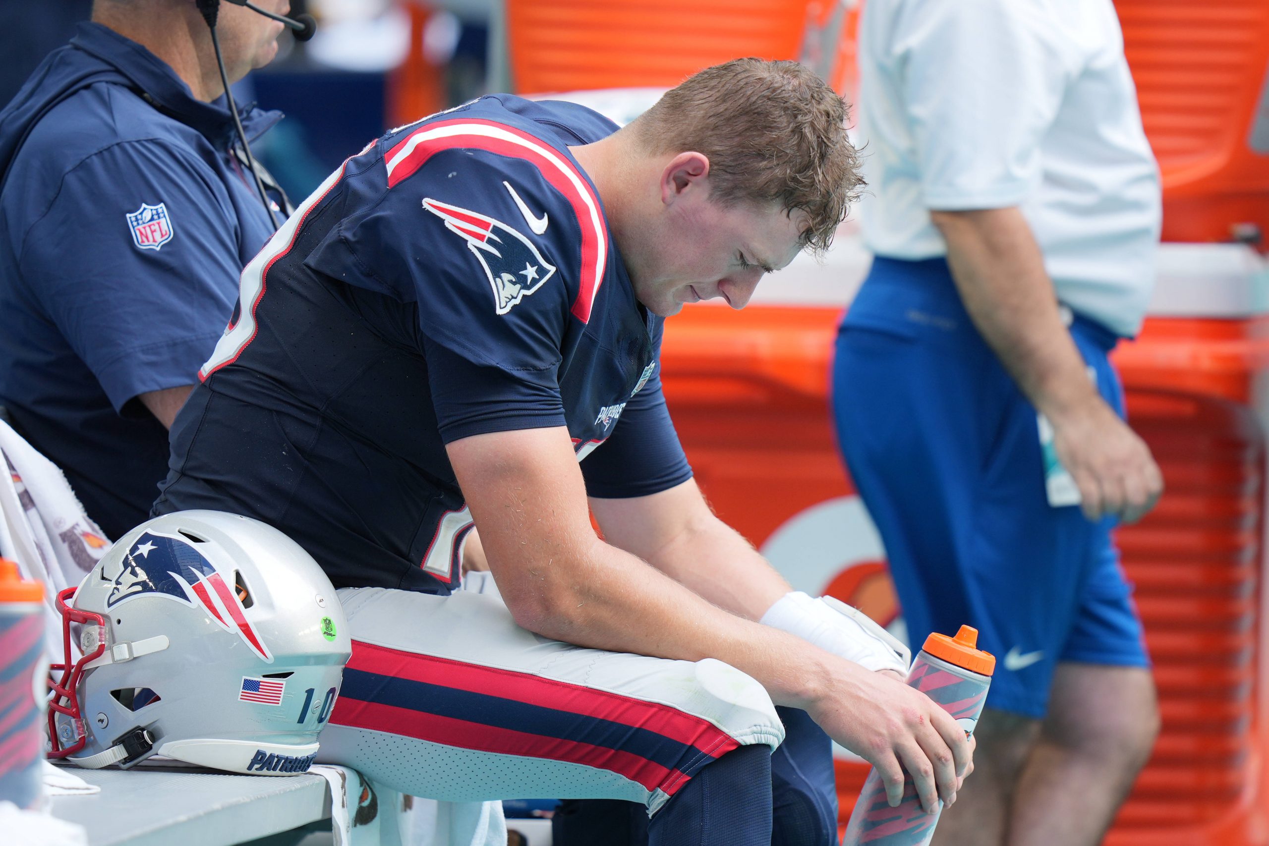 FootballR - NFL - Ein Spieler der New England Patriots, Mac Jones, sitzt ruhig auf der Bank inmitten der Unruhe.