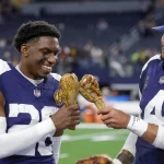 FootballR - NFL Thanksgiving Zuschauerrekord - DaRon Bland und Dak Prescott halten Truthahnkeulen auf einem Feld und erinnern sich dabei an die Geschichte, die durch den aufregenden Pick-6 geschrieben wurde.