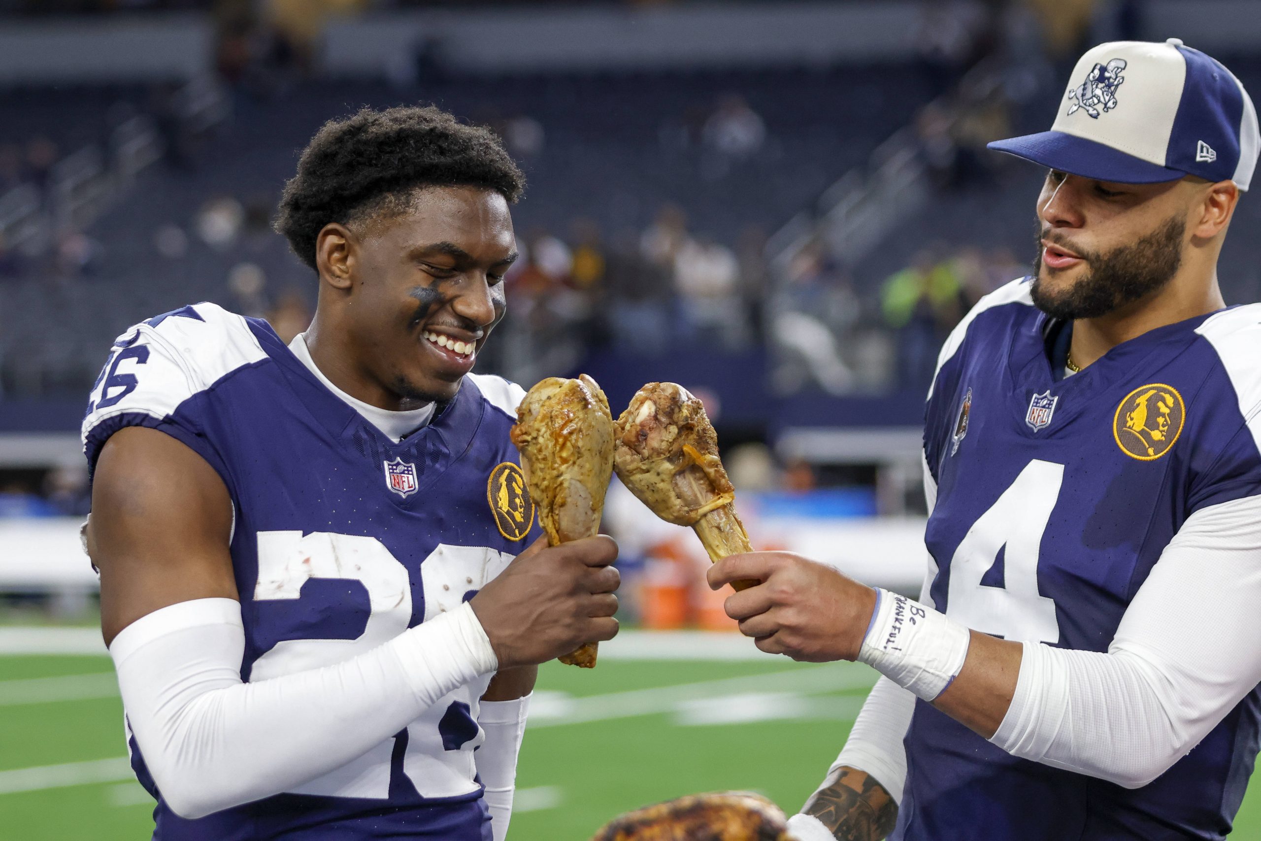 FootballR - NFL Thanksgiving Zuschauerrekord - DaRon Bland und Dak Prescott halten Truthahnkeulen auf einem Feld und erinnern sich dabei an die Geschichte, die durch den aufregenden Pick-6 geschrieben wurde.