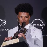 FootballR - NFL - Patriots vor Auswahl eines Quarterbacks - Jayden Daniels hält eine Trophäe vor einer Wand.