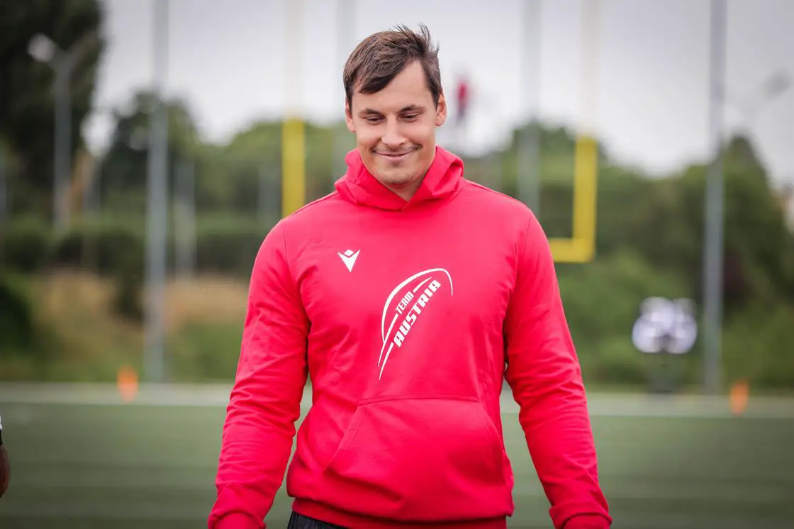 FootballR - NFL - Bernhard Seikovits steht mit einem roten Kapuzenpullover auf einem Footballfeld.