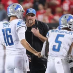 FootballR - NFL - Diese Beschreibung wurde automatisch generiert. Dan Campbell, Cheftrainer der Detroit Lions, spricht während eines Spiels mit seinen Spielern, darunter Jared Goff, und bespricht mit dem Team aggressive Spielstrategien.