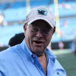 FootballR - NFL - David Tepper, der Besitzer der NFL Carolina Panthers, trägt ein blaues Hemd und eine Mütze und ist auf einem Footballfeld zu sehen.