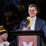 FootballR - NFL - Jim Harbaugh - Ein Mann in Anzug und Krawatte steht an einem Podium und hält eine Rede zum Thema „Gespräche mit den Atlanta Falcons“.