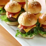 FootballR - NFL - Diese Beschreibung wurde automatisch generiert. Burger-Slider, ein Fanfavorit, serviert auf einem weißen Teller mit Zahnstochern, perfekt für Super-Bowl-Party-Fingerfood.
