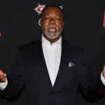 FootballR - NFL - Diese Beschreibung wurde automatisch generiert. Ein Hollywood-NFL-Spieler, Carl Weathers, in einem Anzug mit erhobenen Händen.