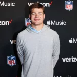 FootballR - NFL - Diese Beschreibung wurde automatisch generiert. Ein Mann in einem grauen Sweatshirt, Drake Maye, steht vor einem NFL-Logo, das möglicherweise auf seine Zukunft als Quarterback hindeutet.