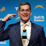 FootballR - NFL Head Coaches - Diese Beschreibung wurde automatisch generiert. Jim Harbaugh, der Cheftrainer der Chargers, zeigt in Anzug und Krawatte den Daumen nach oben.