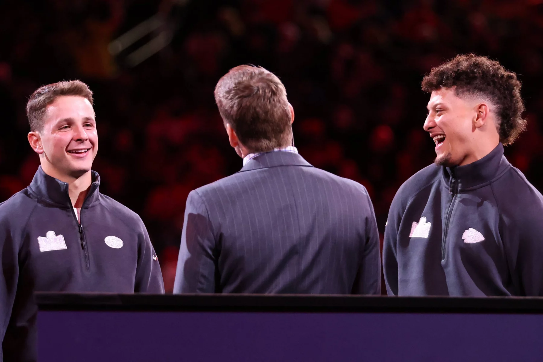 FootballR - NFL 2024 - Diese Beschreibung wurde automatisch generiert. Drei Männer in Anzügen, Brock Purdy, Patrick Mahomes, Scott Hanson unterhalten sich bei der Opening Night während des Super Bowl LVIII.