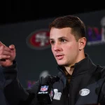 FootballR - NFL - Diese Beschreibung wurde automatisch generiert. Ein Spieler der San Francisco 49ers, Brock Purdy, zeigt während einer Super-Bowl-Pressekonferenz als Starting-QB in die Kamera.