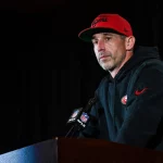 FootballR - NFL - Diese Beschreibung wurde automatisch generiert. Kyle Shanahan, der einen roten Hut trägt, liefert auf dem Podium der San Francisco 49ers einen tollen Auftritt ab.