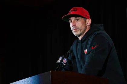 FootballR - NFL - Diese Beschreibung wurde automatisch generiert. Kyle Shanahan, der einen roten Hut trägt, liefert auf dem Podium der San Francisco 49ers einen tollen Auftritt ab.