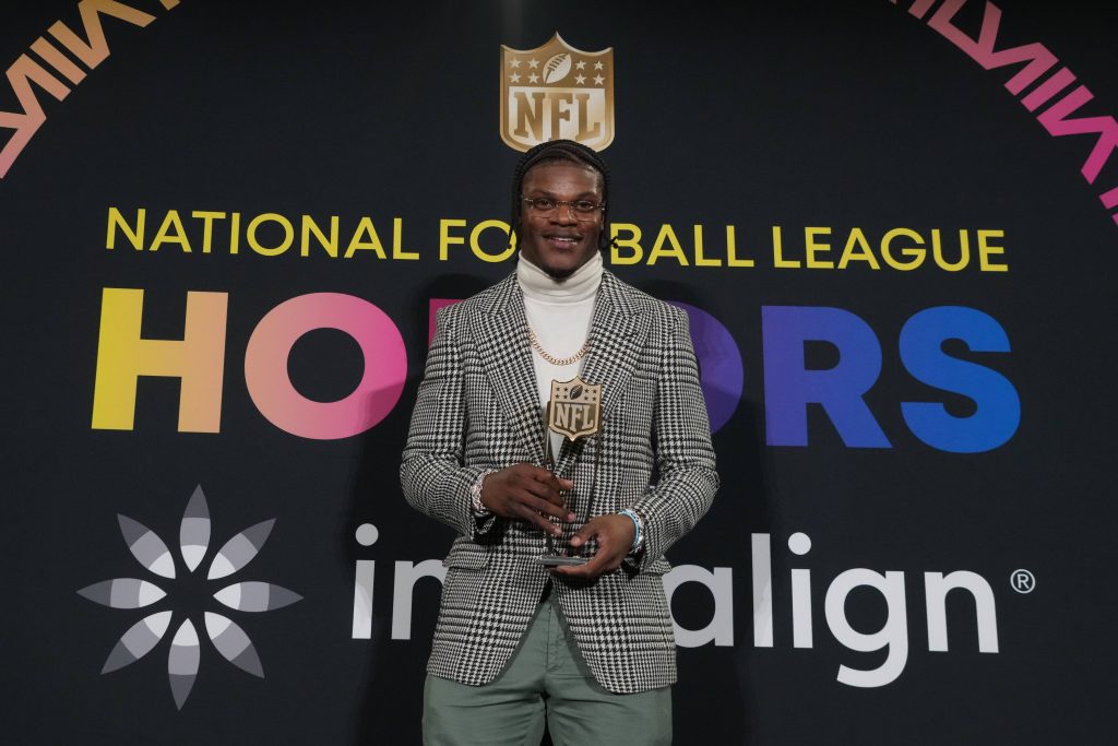 FootballR - NFL MVP Rennen 2024 - Diese Beschreibung wurde automatisch generiert. Lamar Jackson, der NFL MVP 2023, hält stolz seine Auszeichnung für seine bemerkenswerte Leistung in der National Football League.