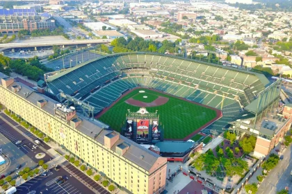 FootballR - NFL - Eine Luftaufnahme zeigt ein sonnendurchflutetes Stadion, umgeben von städtischen Strukturen, mit klarem Himmel darüber und einer geschäftigen Stadtlandschaft dahinter, die die Fußballkultur hervorhebt. Diese Beschreibung wurde automatisch generiert.