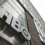 FootballR - NFL - Eine flache Ansicht der Fassade eines modernen Gebäudes mit dem auffälligen, dreidimensionalen „HBO“-Logo, das deutlich vor dem eleganten Außendesign zu sehen ist und die Präsenz des Unternehmens und die Vorfreude auf Hard Knocks symbolisiert Diese Beschreibung wurde automatisch generiert.