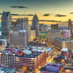 FootballR - NFL - Ein Panoramablick auf eine geschäftige Stadtlandschaft in der Abenddämmerung, mit Wolkenkratzern, die in die warmen Farben des Sonnenuntergangs getaucht sind, und Stadtlichtern, die am Abendhimmel zu funkeln beginnen, in sehnsüchtiger Vorfreude auf den NFL Draft Diese Beschreibung wurde automatisch generiert.