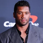 FootballR - NFL - Diese Beschreibung wurde automatisch generiert. Russell Wilson - Ein Mann in einem grauen Anzug steht auf einem roten Teppich bei einer Broncos-Veranstaltung.