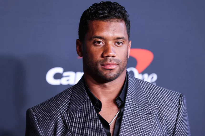 FootballR - NFL - Diese Beschreibung wurde automatisch generiert. Russell Wilson - Ein Mann in einem grauen Anzug steht auf einem roten Teppich bei einer Broncos-Veranstaltung.