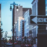 FootballR - NFL - Ein Einbahnstraßenschild vor einem städtischen Hintergrund mit verschwommenen Gebäuden und Straßenlaternen, das die Essenz des Stadtlebens in der Abenddämmerung einfängt. Diese Beschreibung wurde automatisch generiert.