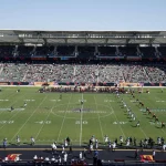 FootballR - NFL Regeländerungen - Kickoff - In einem Stadion findet gerade ein American-Football-Spiel statt, die Mannschaften stehen für den Anpfiff auf dem Spielfeld aufgereiht und die Tribünen sind unter strahlend blauem Himmel mit Zuschauern gefüllt. Diese Beschreibung wurde automatisch generiert.