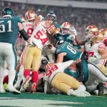 FootballR - NFL - Ein dynamischer Moment während eines American-Football-Spiels, in dem Spieler zweier Teams in einem Gewirr aus Gliedmaßen und Ausrüstung aufeinanderprallen, um die Vorherrschaft auf dem Spielfeld kämpfen und ein Tush Push Manöver ausführen. Diese Beschreibung wurde automatisch generiert.