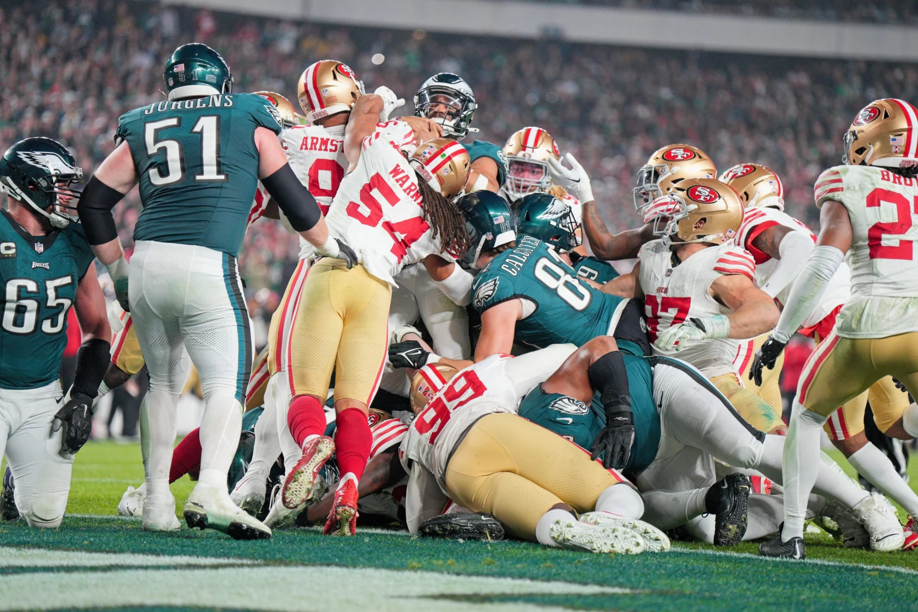 FootballR - NFL - Ein dynamischer Moment während eines American-Football-Spiels, in dem Spieler zweier Teams in einem Gewirr aus Gliedmaßen und Ausrüstung aufeinanderprallen, um die Vorherrschaft auf dem Spielfeld kämpfen und ein Tush Push Manöver ausführen. Diese Beschreibung wurde automatisch generiert.