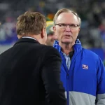 FootballR - NFL - Zwei Männer im Gespräch auf einem Footballplatz; Einer trug eine blau-rote Jacke und Brille der New York Giants, identifiziert als John Mara, der andere trug eine dunkle Jacke, mit verschwommenen Zuschauern im Bild Diese Beschreibung wurde automatisch generiert.