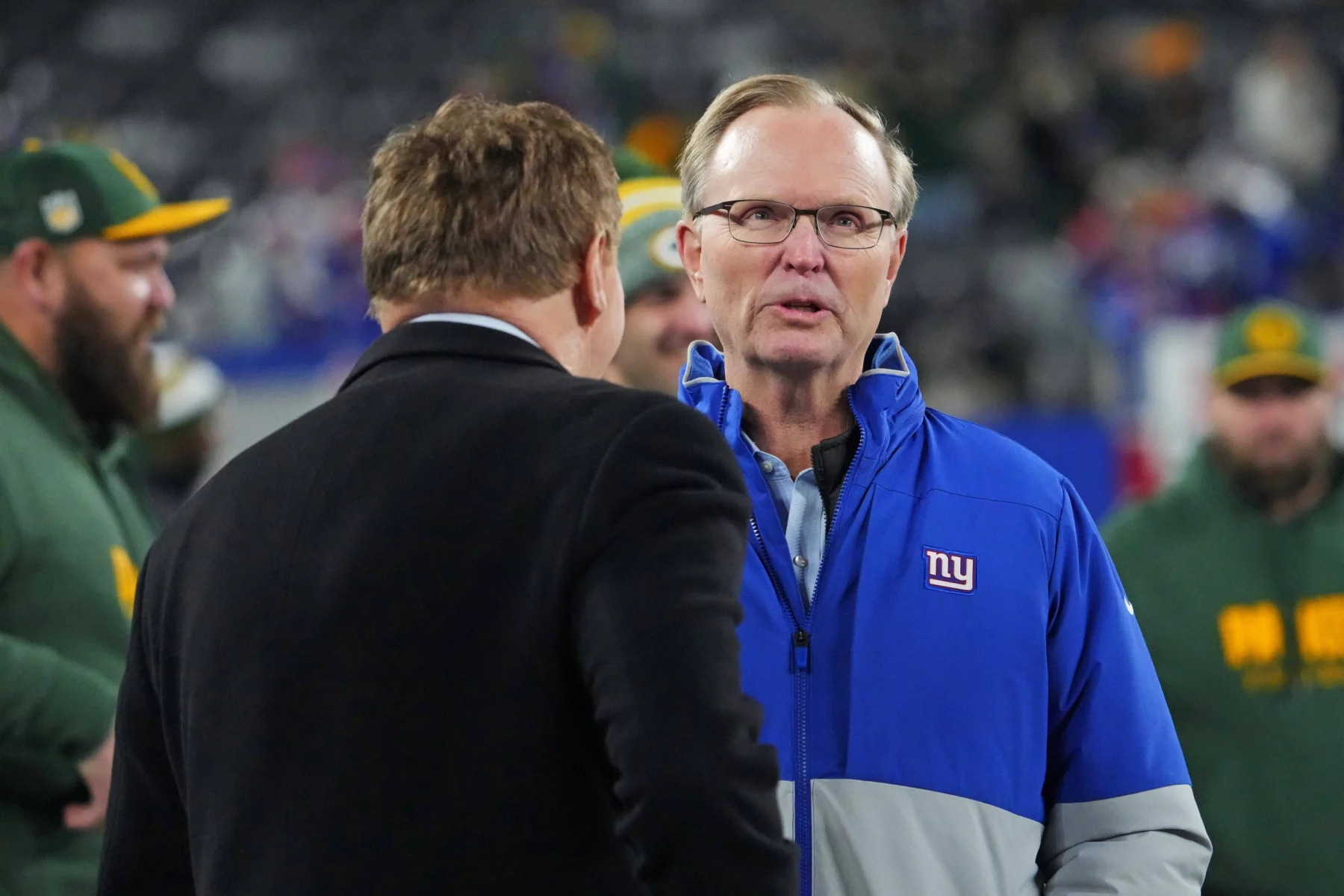 FootballR - NFL - Zwei Männer im Gespräch auf einem Footballplatz; Einer trug eine blau-rote Jacke und Brille der New York Giants, identifiziert als John Mara, der andere trug eine dunkle Jacke, mit verschwommenen Zuschauern im Bild Diese Beschreibung wurde automatisch generiert.