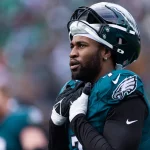 FootballR - NFL - Ein American-Football-Spieler, Haason Reddick, der jetzt die Ausrüstung der New York Jets trägt, posiert aufmerksam auf dem Spielfeld und strahlt vor dem Spiel Konzentration und Entschlossenheit aus. Diese Beschreibung wurde automatisch generiert.
