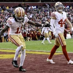 FootballR - NFL - Zwei Footballspieler in weißen und roten 49ers-Uniformen feiern auf dem Spielfeld; einer (Brandon Aiyuk) streckt triumphierend seinen Arm aus, während er den Ball hält, möglicherweise nach einem erfolgreichen Spiel. Diese Beschreibung wurde automatisch generiert.