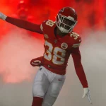 FootballR - NFL - L'Jarius Sneed - Ein Footballspieler, der ein rot-goldenes Trikot mit der Nummer 38 trägt, feiert auf dem Spielfeld mit ausgestrecktem Arm vor einer Kulisse aus dramatischem rotem Rauch. Diese Beschreibung wurde automatisch generiert.