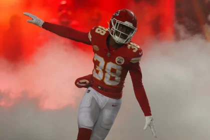 FootballR - NFL - L'Jarius Sneed - Ein Footballspieler, der ein rot-goldenes Trikot mit der Nummer 38 trägt, feiert auf dem Spielfeld mit ausgestrecktem Arm vor einer Kulisse aus dramatischem rotem Rauch. Diese Beschreibung wurde automatisch generiert.