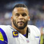 FootballR - NFL - Ein konzentrierter American-Football-Spieler, der ein weiß-blaues Rams-Trikot mit dem „C“-Aufnäher des Kapitäns trägt, hört abseits des Spielfelds aufmerksam zu und demonstriert Entschlossenheit und Führung inmitten der Intensität eines Spiels. Aaron Donald. Diese Beschreibung wurde automatisch generiert.