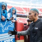FootballR - NFL - Diese Beschreibung wurde automatisch generiert. Ein Mann mit blauem Hut unterhält sich mit Fans bei einem Footballspiel der Detroit Lions. Amon-Ra St. Brown