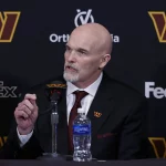 FootballR - NFL - Dan Quinn im Anzug mit Krawatte mit dem markanten Logo der Washington Commanders sitzt auf einer Pressekonferenz und wendet sich selbstbewusst an das Publikum, im Hintergrund sind Sponsorenbanner zu sehen. Diese Beschreibung wurde automatisch generiert.