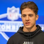 FootballR - NFL - Diese Beschreibung wurde automatisch generiert. Ein junger Wide Receiver, Luke McCaffrey, in einem schwarzen Kapuzenpullover steht vor einem NFL-Logo.