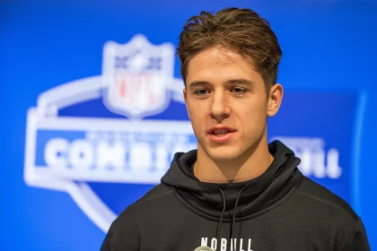 FootballR - NFL - Diese Beschreibung wurde automatisch generiert. Ein junger Wide Receiver, Luke McCaffrey, in einem schwarzen Kapuzenpullover steht vor einem NFL-Logo.