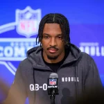 FootballR - NFL - Ein konzentrierter Athlet, Jayden Daniels, bei einem Pro Day, der ein T-Shirt mit QB-Thema und das Logo eines Sponsors trug, nahm an einer Medieninteraktion unter hellem Licht vor dem Hintergrund des Liga-Brandings teil. Diese Beschreibung wurde automatisch generiert.